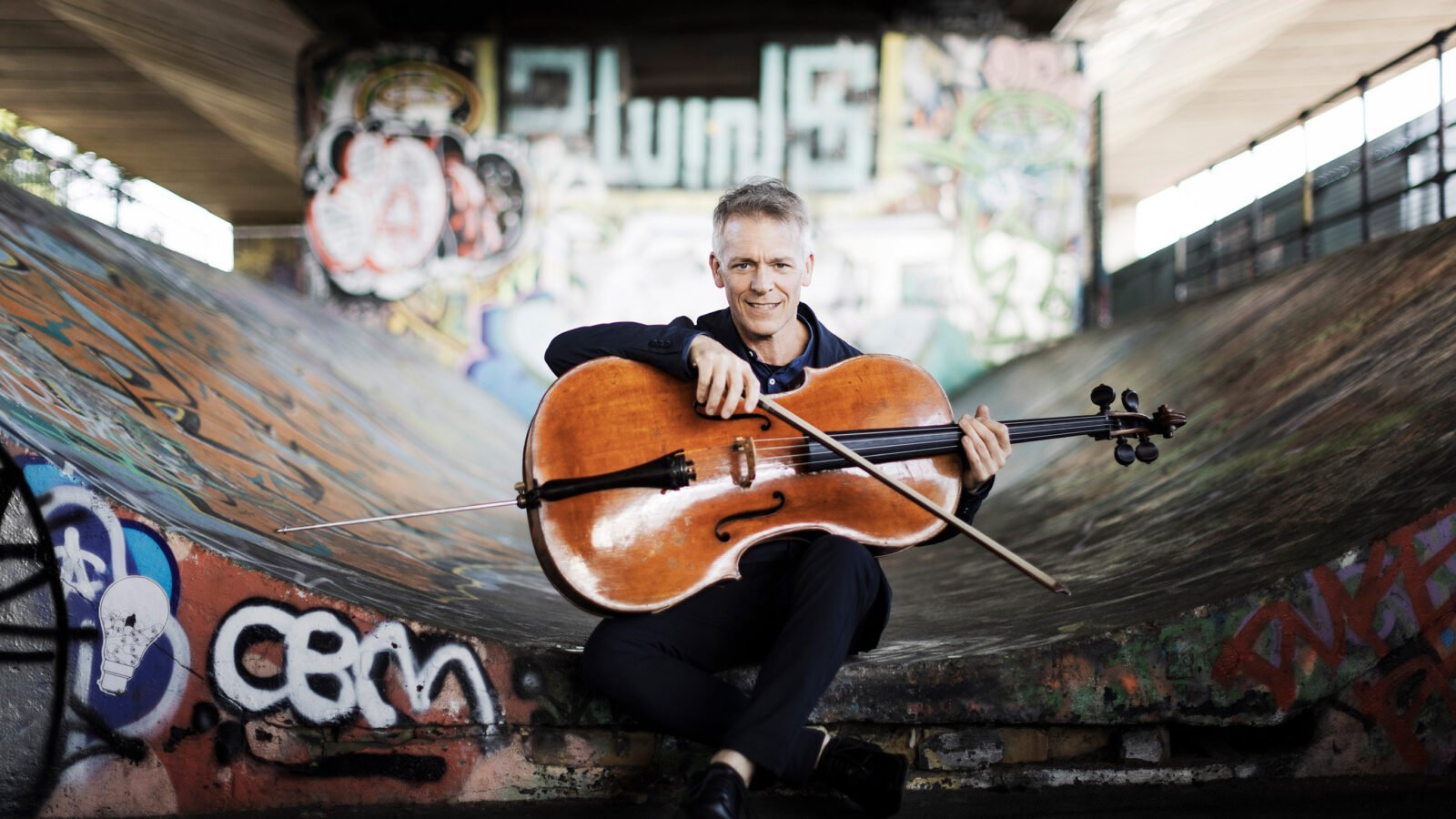 Alban Gerhardt smiling holding a cello against a graffiti-laden urban backdrop