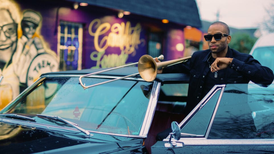Trombone Shorty rests his instrument on a car with a colorful mural in the background